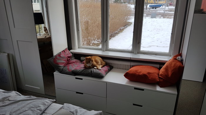 Sovrum med vit byrå, en rödhårig katt som vilar på en kudde vid fönstret med utsikt över snötäckt mark.