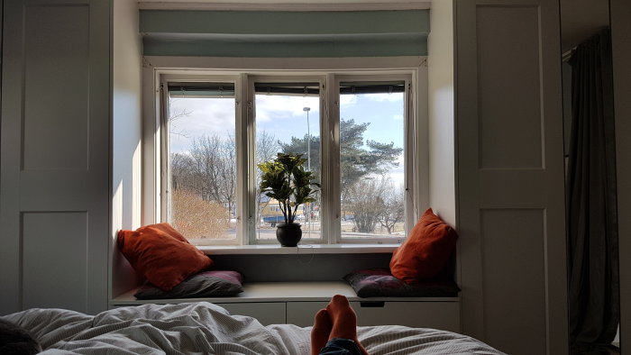 Vy från sovrummet med blick mot fönstret, kuddar på fönsterbrädan och personens fötter i förgrunden.