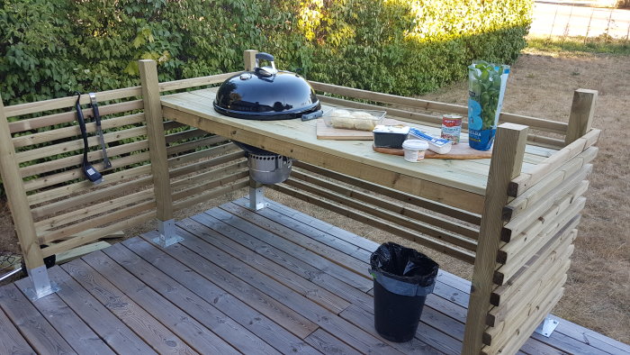 Nybyggt utomhuskök med grill och matlagningsutrustning på en träterrass och nysatt häck i bakgrunden.