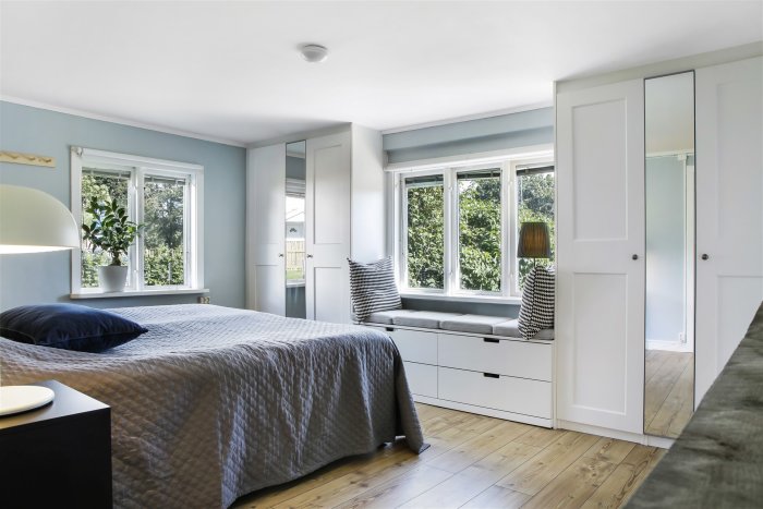 Stilrent sovrum med grå säng, inbyggda skåp och fönstersits mot grönska.