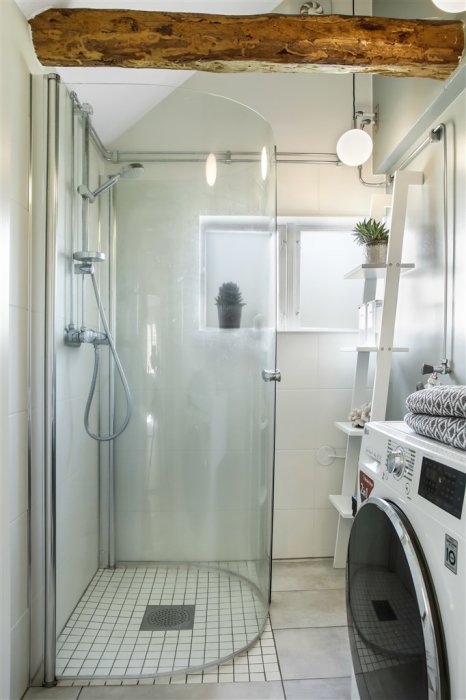 Renoverat badrum med duschhörna, träbalk i taket, vit tvättmaskin och hyllor med växter.