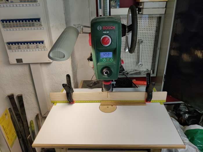 DIY borrställ med laminerad kökslucka som bord, vinkelprofil, tvingar och pelarborr med mätverktyg.