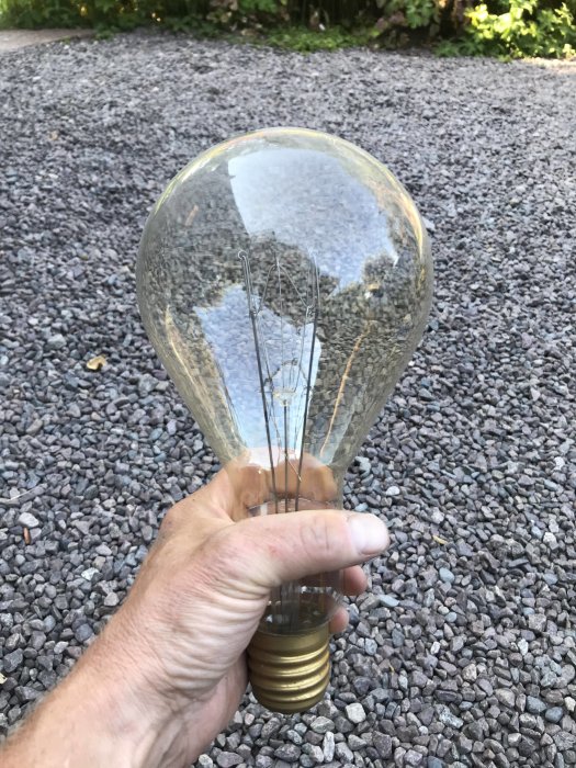 Hand håller en stor glödlampa på grusunderlag, troligen E40 sockel för byggstrålkastare.