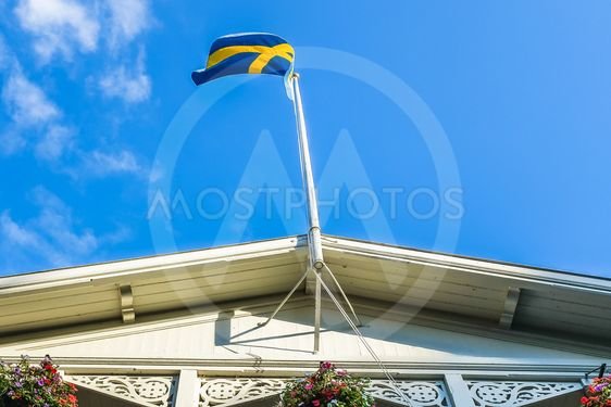 Svensk flagga på en flaggstång monterad ovanpå ett vitt hus med dekorativa träsnitt och blomlådor.