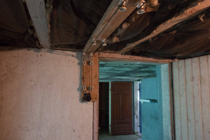 Källarutrymme med öppen dörr och bar elinstallation i taket med synliga träbjälkar och isolering.