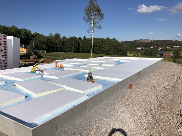 Grundisolering med plast och frigolit på byggplats under solig himmel med natur och hus i bakgrunden.