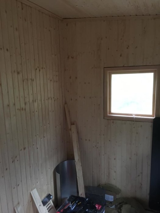 Hörn av ett rum under renovering med träpanel på väggarna, ett fönster och byggmaterial på golvet.