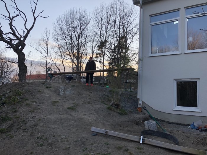 Person som står vid en pågående altanbyggnation bredvid hus med redskap och byggmaterial i förgrunden.