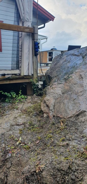 En hörna av ett hus med eluttag på en trästolpe och en stor sten i förgrunden som del av ett byggprojekt.
