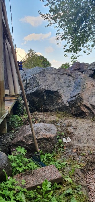 Hörn av en byggnad med en blå vattenslang, stora stenblock och en kratta på en ojämn mark täckt med gräs och löv.
