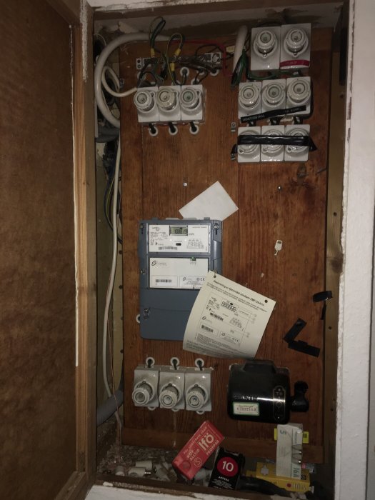 Öppen elcentral med säkringar, mätare och ledningar på en trävägg, oordning runtomkring.