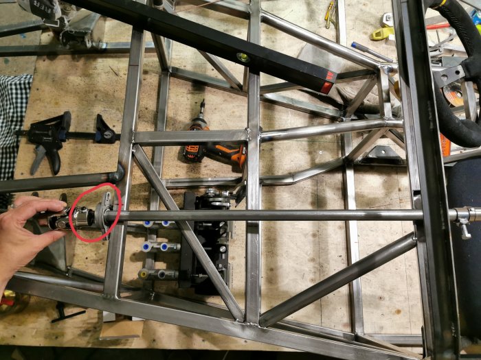 Hand som pekar på en knut på en bilaxel inuti en metallram i ett garage, med verktyg utspridda i bakgrunden.