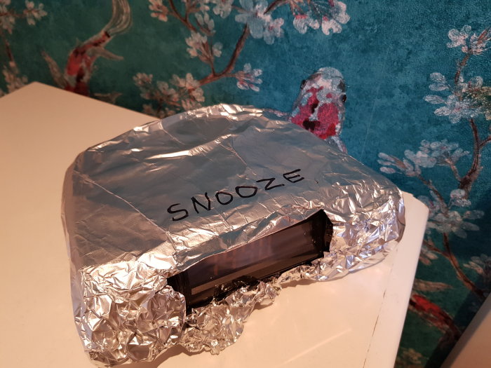 Klockradio täckt med aluminiumfolie och ordet "SNOOZE" skrivet på toppen, belägen på ett vitt underlag mot tapet med blommönster.