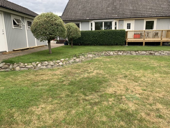 Trädgård med slänt mark och stenrad, gräsmatta framför hus med veranda och träd.