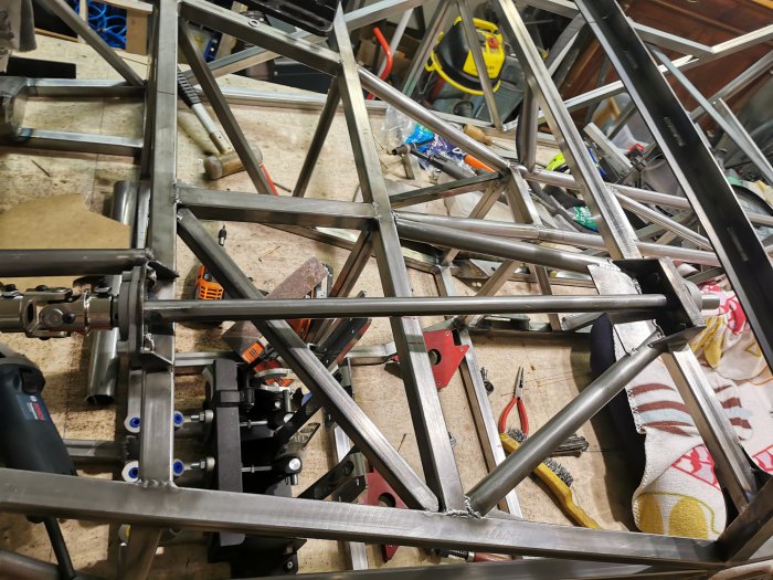Metallkonstruktion med många korsande rör och svetspunkter i ett verkstadsutrymme med verktyg och utrustning i bakgrunden.