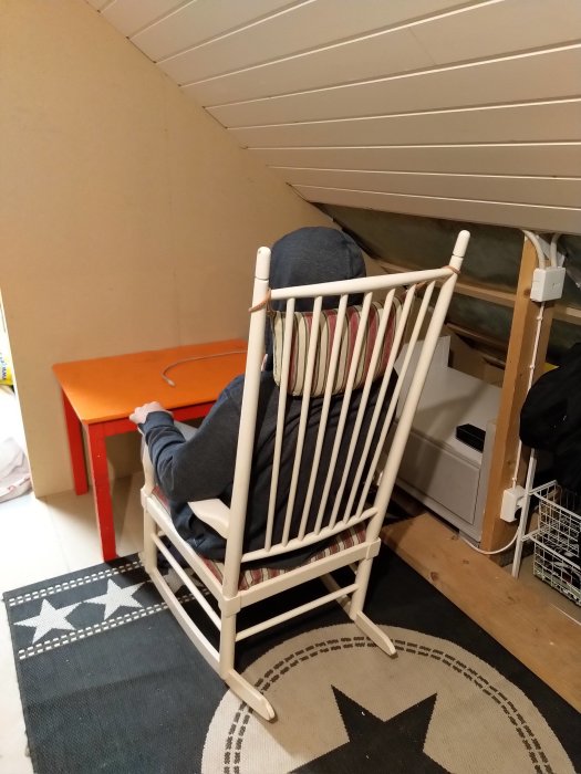 Person sitter i en vit gungstol framför ett orange skrivbord under snedtaket på ett loft.