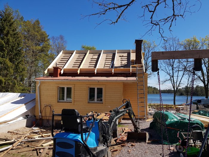 Renovering av en gul sommarstuga från 1958 med nytt takstomme under blå himmel, byggmaterial och maskiner syns framför.