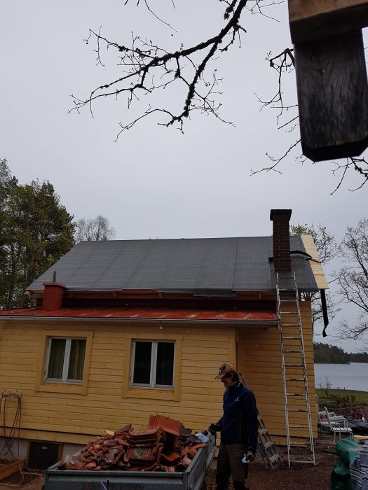 Renovering av gammal gul sommarstuga med person och material framför, omgiven av natur och sjöutsikt.