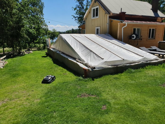 Stommen till ett husbygge täckt med presenning på en gräsmatta, med trähus i bakgrunden under en klar himmel.