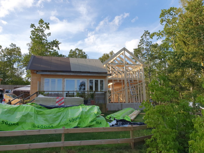 Ny trästomme under konstruktion bredvid befintligt hus, omgivet av grönska och byggmaterial på marken.