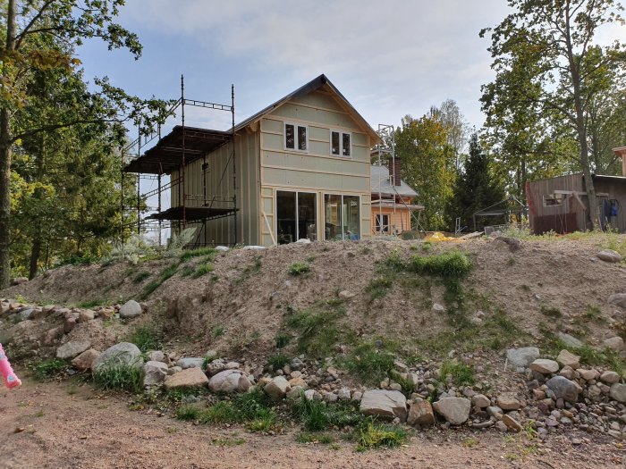 Hus under renovering med isoleringsskivor synliga, byggnadsställning och ojämn terräng med stenar.