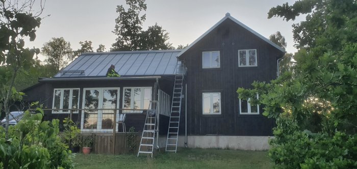 Hus med svartbrun fasad, person på taket vid solnedgång, och stege lutad mot huset.