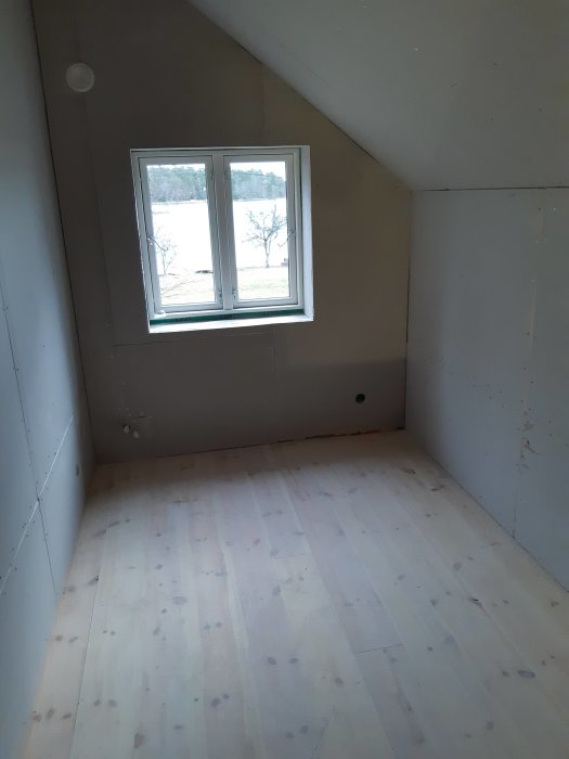 Interiör av ett rum under renovering med nygipsade väggar och obehandlat trägolv med ett fönster.