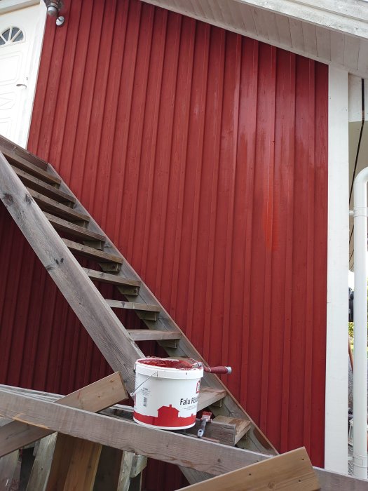 Hink med rödfärg på en trästege framför en nyss målad röd träfasad på ett garage.