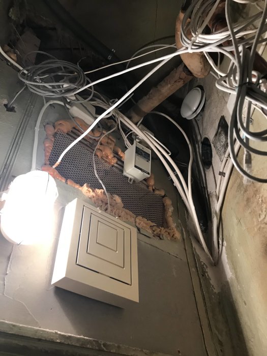 Oorganiserad installation av rör och kablar i hörn av ett rum med klinkergolv och råa väggar.