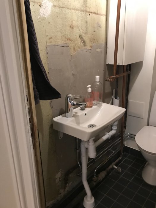 Orenoverat badrum med handfat, toalett och synliga rör mot slitna oavslutade väggar och klinkergolv.