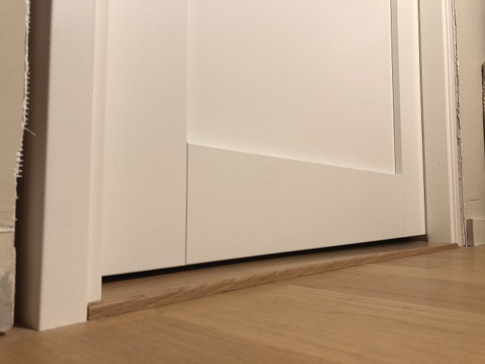 Ojämnt mellanrum under vit dörr och trägolv, högre tröskel på ena sidan.