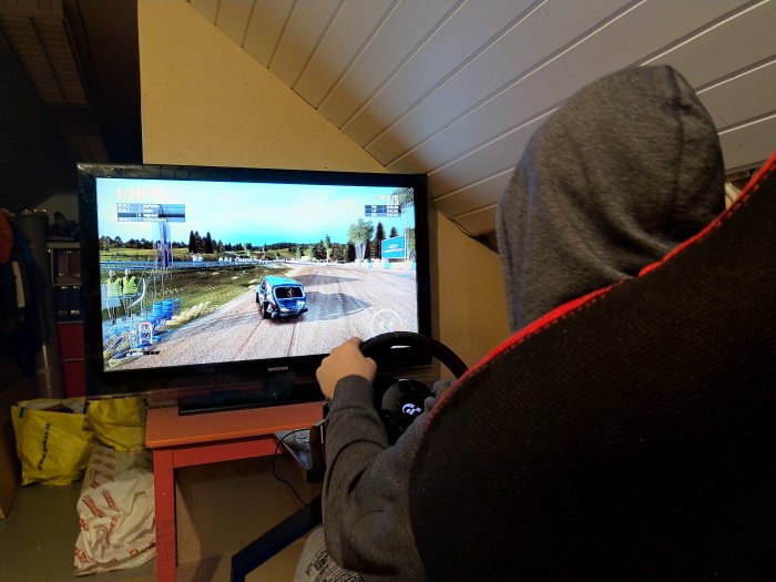 Person spelar racingspel med ratt och pedaler framför TV i hörnan av ett rum.