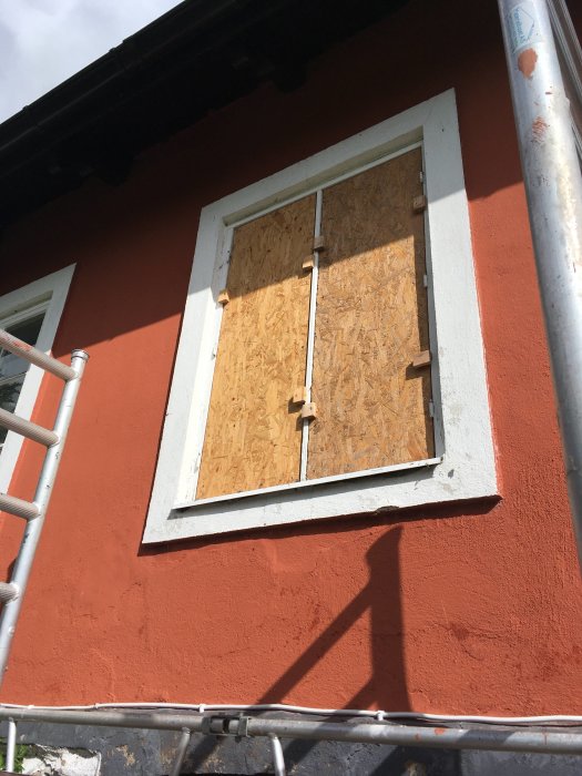 Fönster utan glas med falska bågar och OSB-skivor monterade i en röd husvägg.
