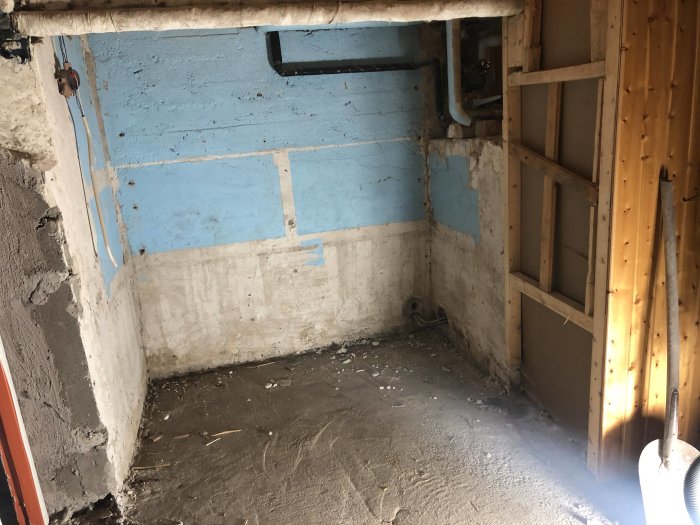 Hörn av ett rum under renovering med blottlagda plåtreglar, byggplywood och exponerade rör.