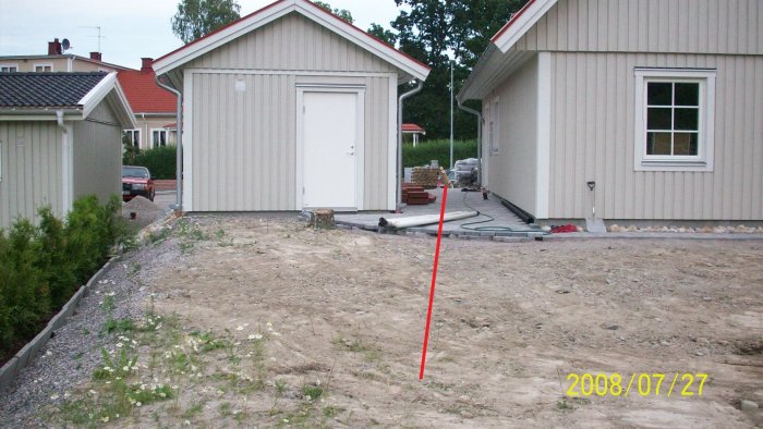 Gräsmark mellan husgrund och förråd med synliga VVS-installationer, indikerad grävningslinje markerad i rött.