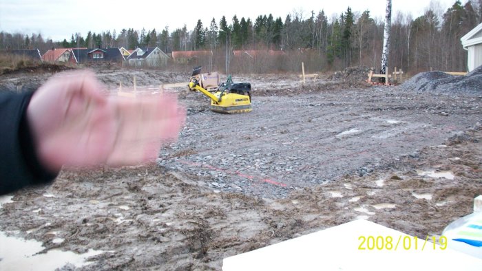 Byggplats med grävmaskin mellan husgrunden och förråd, ogynnsamma arbetsförhållanden.