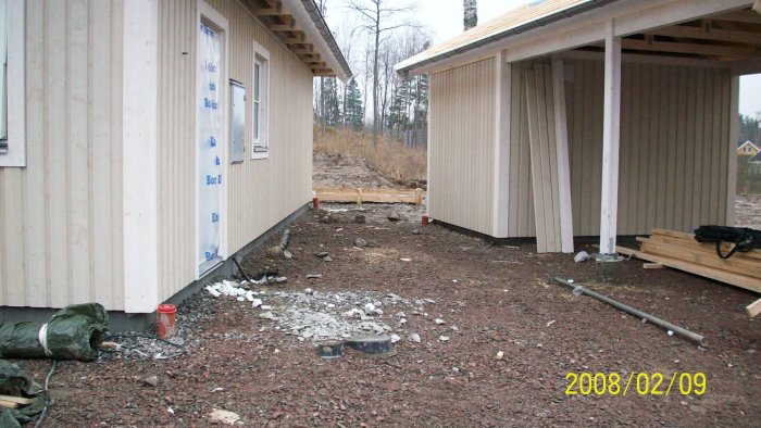 Trång byggarbetsplats mellan hus och förråd med början på grävarbete och byggmaterial på marken.