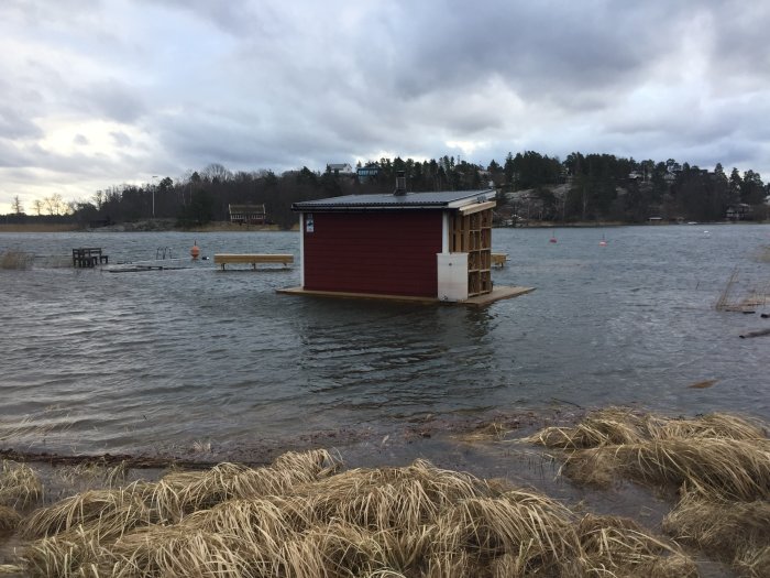 Röd bastubyggnad nära vattnets kant med omgivande högt vatten, bastun stöds av tillfälliga domkrafter.