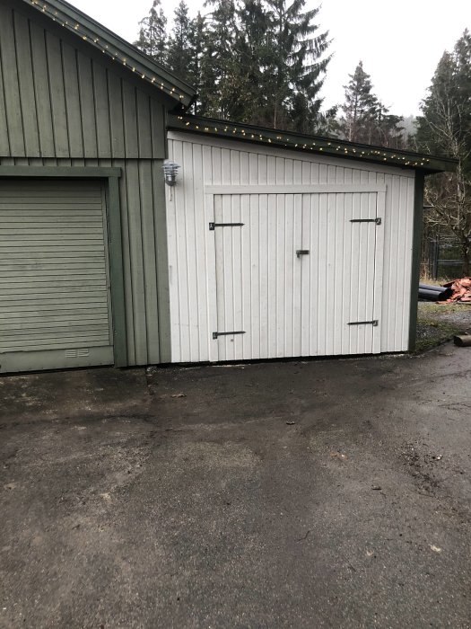 Vit baspanel från Byggmax på en förrådsbyggnad med dubbla dörrar, bredvid ett grönt garage.