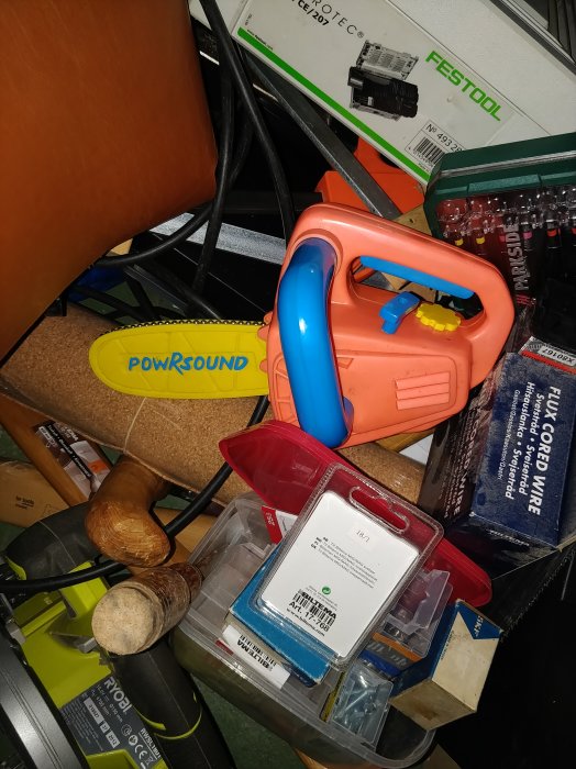 Ett rörigt arbetsbord med en barnleksaksmotorsåg, verktyg och diverse byggmaterial i en oorganiserad hög.