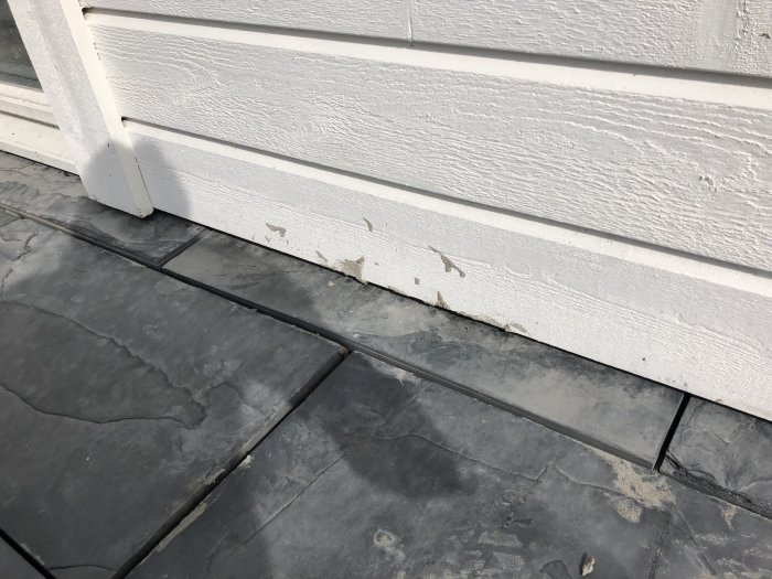 Skadad vit träpanel nära marken med grå stenplattor som ligger nära utan tydligt mellanrum.