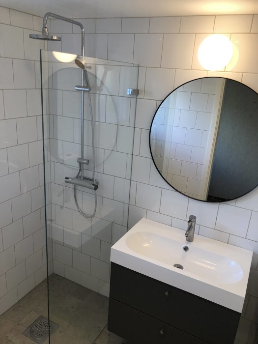 Nytt badrum med glasduschvägg, vit handfat på svart kommod, rund spegel och vägglampa.