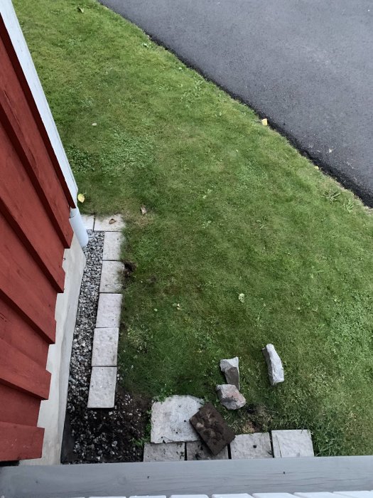 Bild av väg mot hus med trottoarkant, gräsmatta och stenplattor som leder vatten mot husets grund.