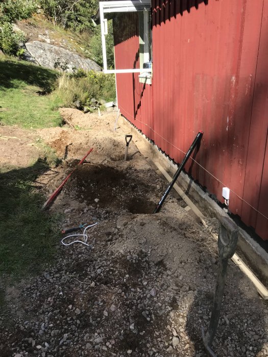 Uppgrävningsarbete vid husvägg med redskap, rör för avrinning och synligt berg.