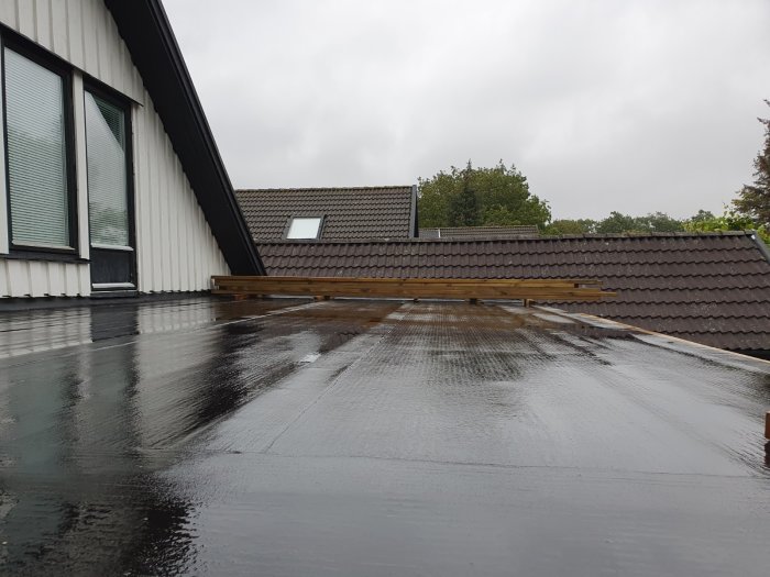 Ett tak med låg lutning, täckt av en svart derbigum-matta, med reglar som förbereds för trall intill ett hus.