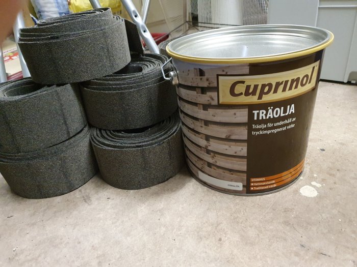 En burk med träolja märkt Cuprinol och högar av svarta derbigum-remsor på ett betonggolv.