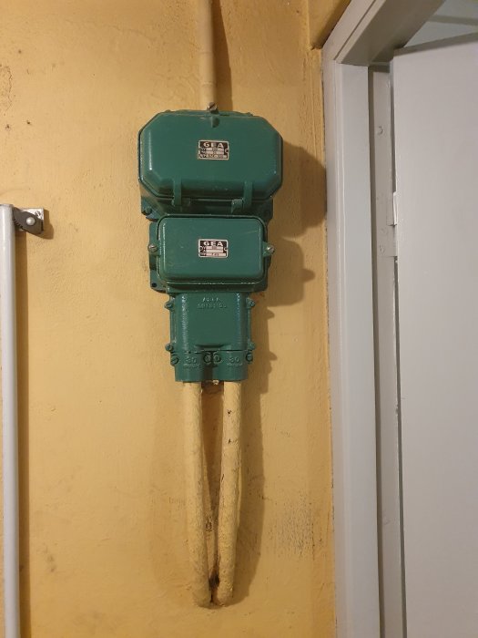 Två gröna elektricitetsmätare monterade ovanför varandra på en gul vägg intill en vit dörr.