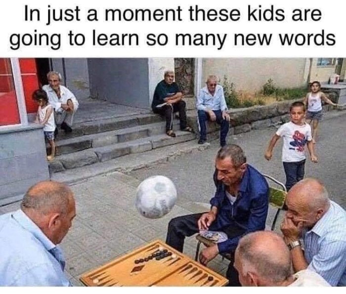 Barn springer mot boll nära män som spelar backgammon på trottoar, åskådare i bakgrunden.