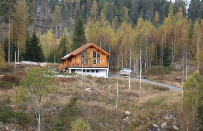 Ett nybyggt träfritidshus i en skogsglänta med släpvagn parkerad intill en grusväg.