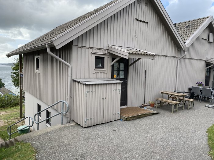 En grå förrådsbod med vita snickeridetaljer placerad bredvid en byggnad med liknande exteriör och takvinkel.